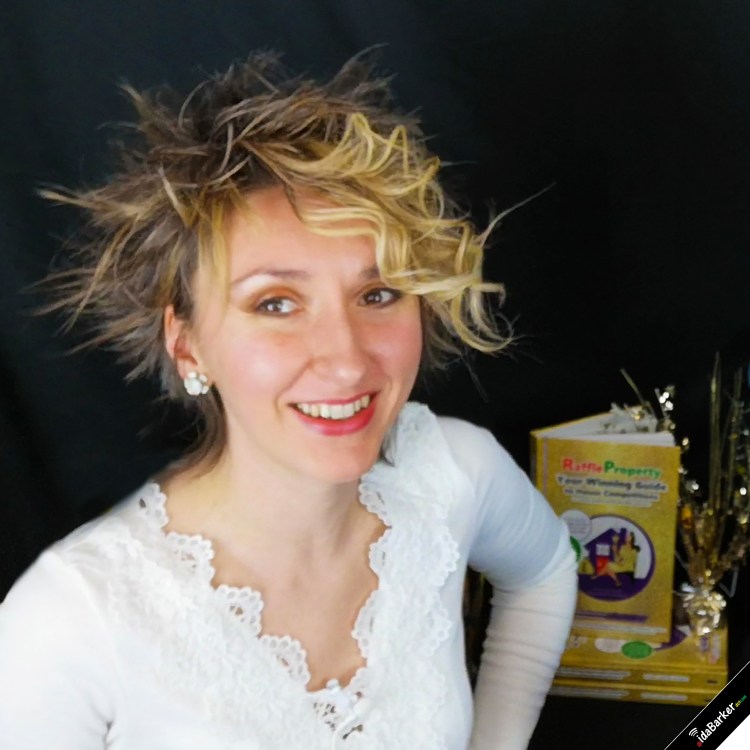 Profile photo taken in 2021 of Ida Barker; wearing a white lace long-sleeve top; she is openly smiling; with curly bouncy hair at the front of her head; in the background we can see her co-authored guidebook, "Raffle Property: Your Winning Guide to House Competitions (for entrants, property-owners and charity organisers)"