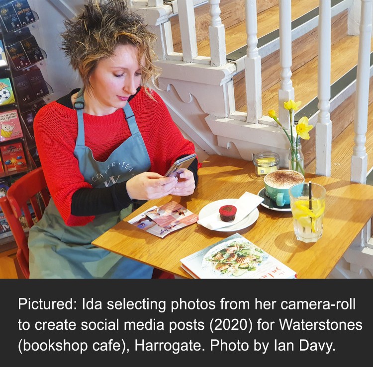 Image shows Ida Barker sat at a table with a cup of coffee and cake, she is concentrating on looking at her mobile phone, selecting photos from her camera-roll to create social media posts (in 2020) for Waterstones (bookshop cafe), Harrogate. Photo by Ian Davy.
