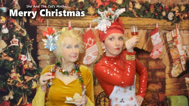 She And The Cat's Mother (mother and daughter, Gwen Hullah and Ida Barker) wishing you a very Merry Christmas; they are looking festive beside a fireplace decorated with Christmas tinsel and surrounded by festivities.